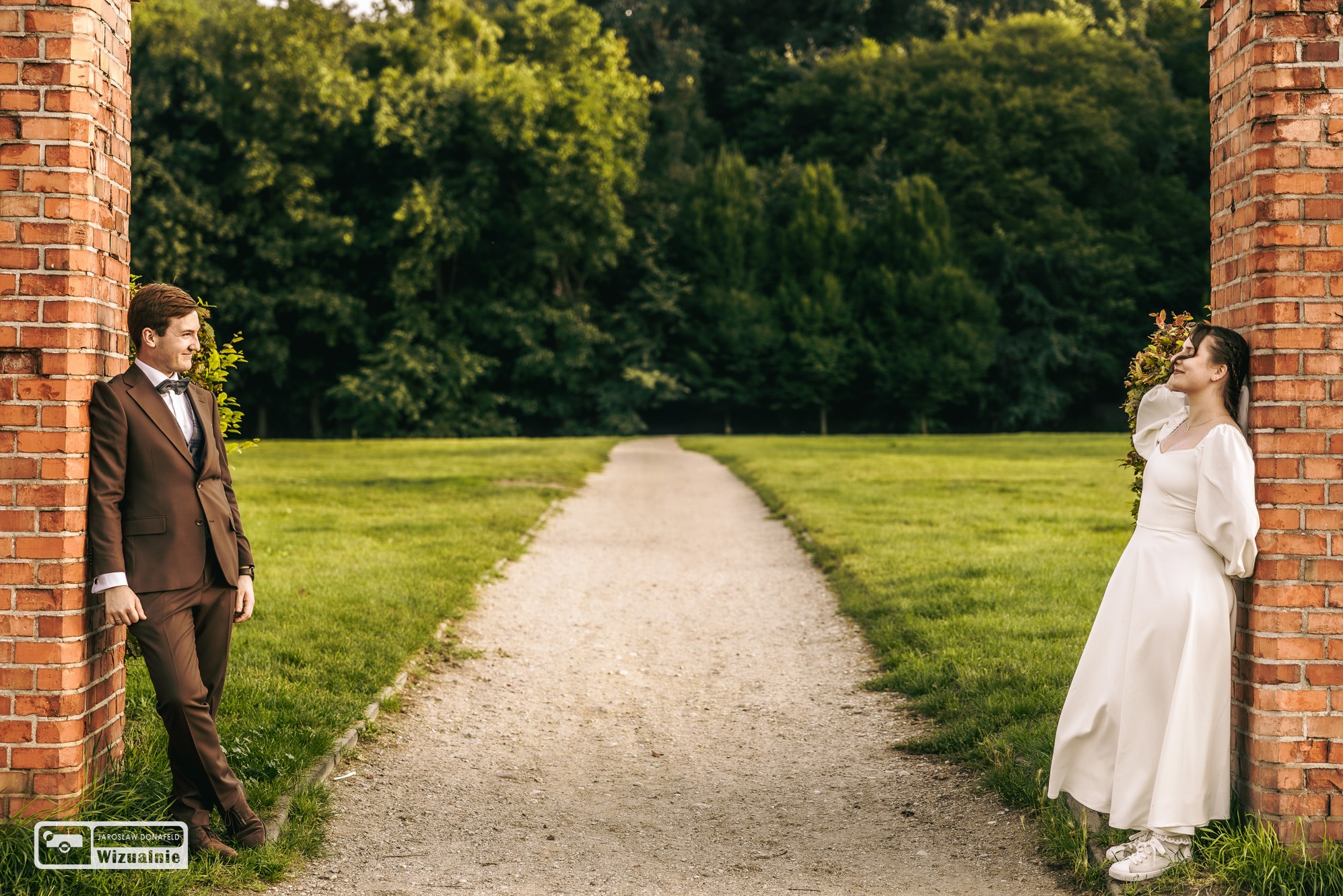 Para narzeczeńska w parku: mężczyzna w brązowym garniturze oparty o ceglany filar, kobieta w białej sukience i trampkach oparta o drugi filar, na tle zieleni i ścieżki.