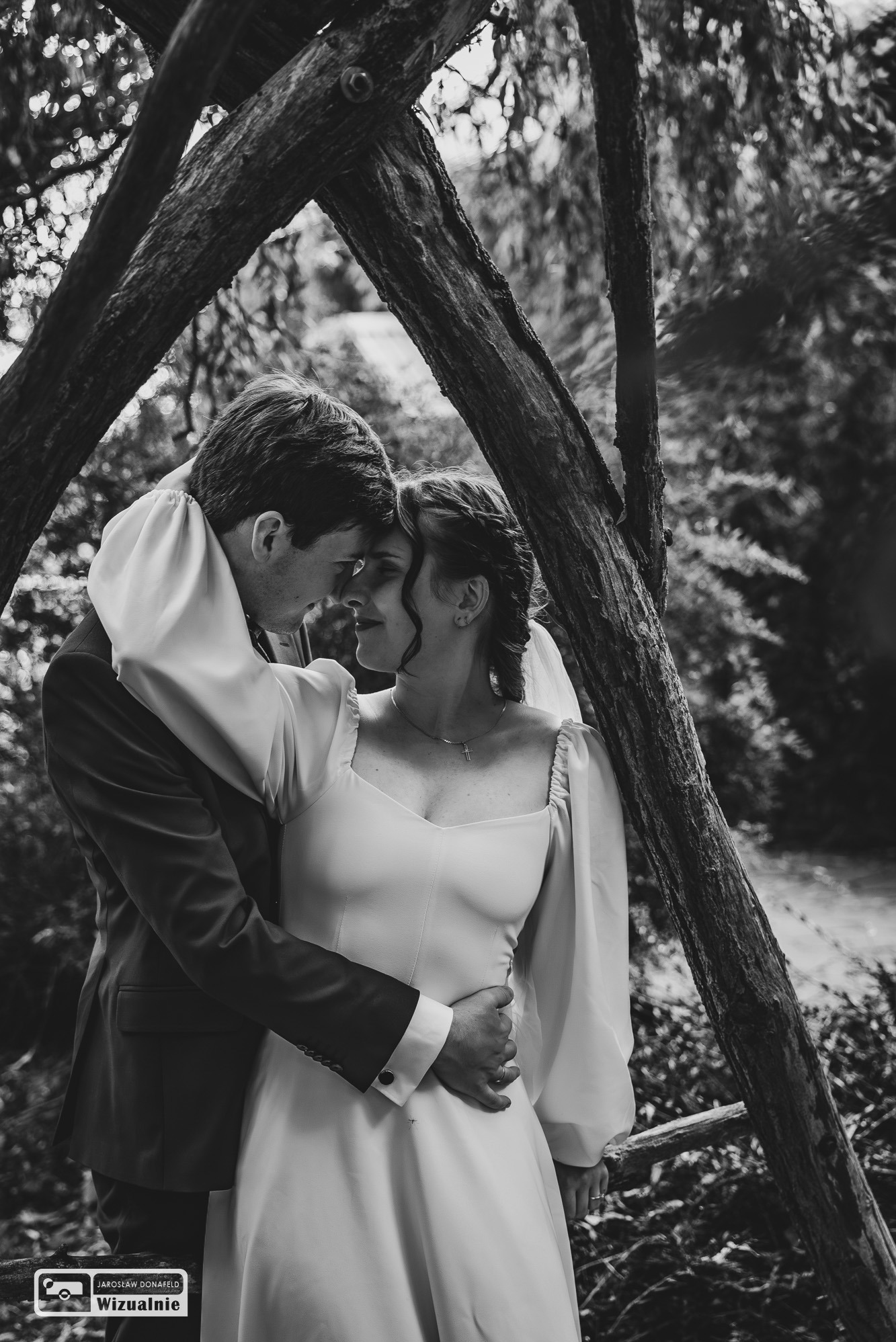 Czarno-białe zdjęcie pary młodej w objęciach, z bliska, z drzewem w tle, panna młoda w sukni z długimi rękawami i upiętymi włosami.
