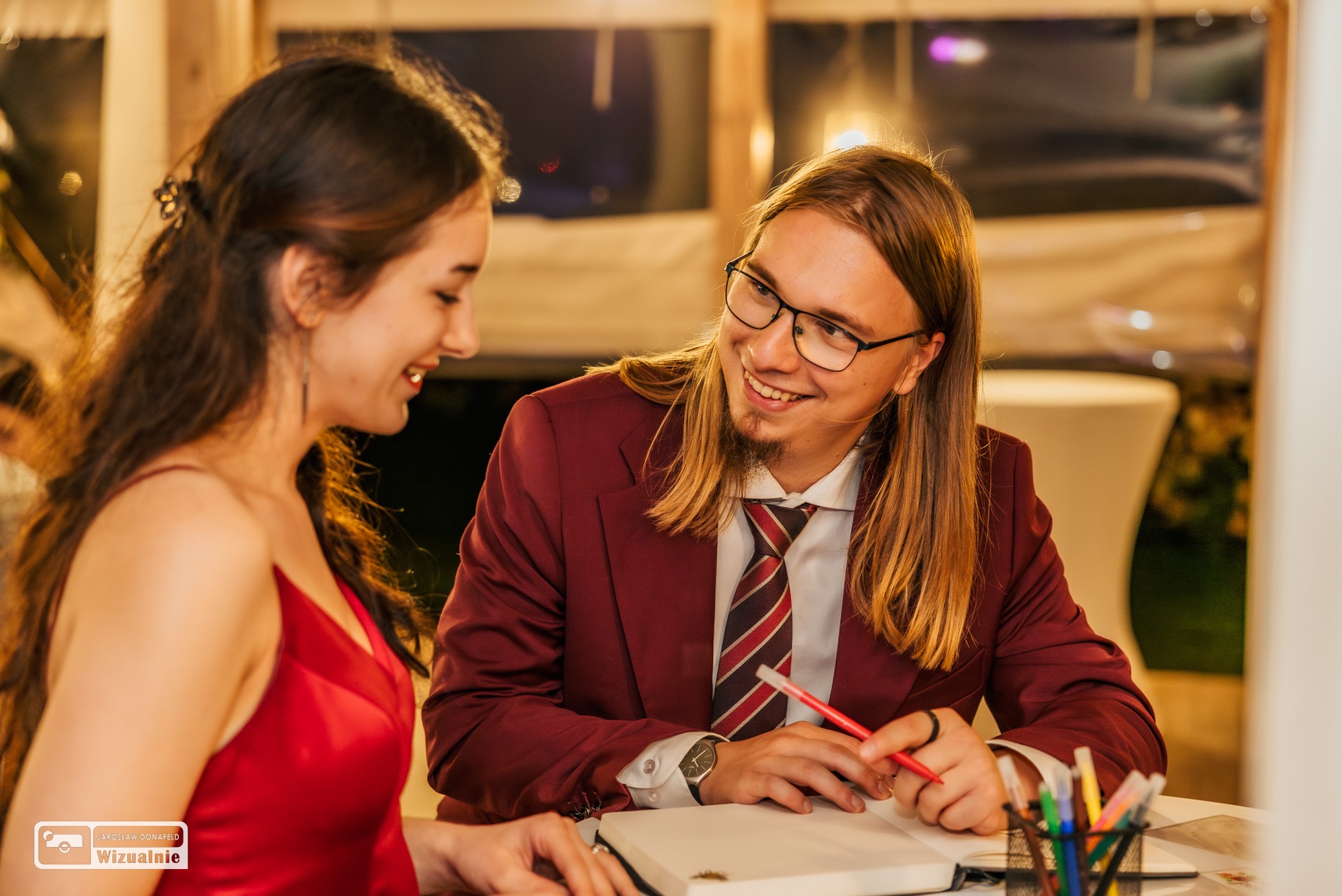 Młoda para w eleganckich strojach, uśmiechnięta i patrząca na siebie podczas planowania, mężczyzna trzyma czerwony ołówek nad otwartym notatnikiem, na stole kolorowe pisaki w metalowym pojemniku.