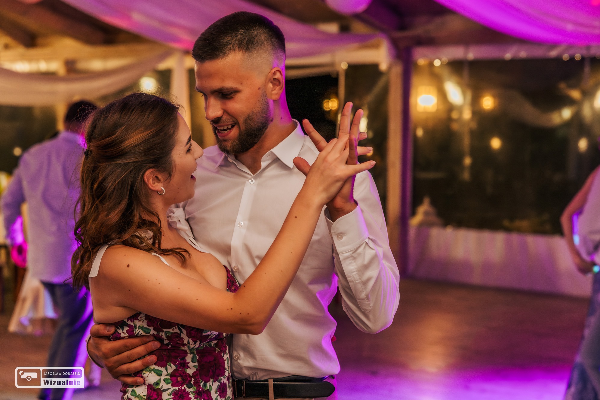 Uśmiechnięta para młoda w tańcu podczas wesela, ona w sukience w kwiaty, on w białej koszuli, oświetlenie sceniczne w tle.