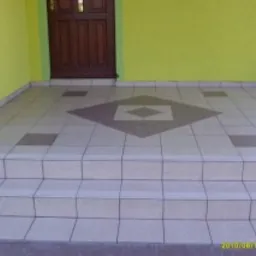 Wyłożony jasnymi płytkami ceramicznymi taras przed wejściem do domu, z dekoracyjnym wzorem rombu w odcieniach brązu na środku, widoczne schody z płytek i fragment ściany w kolorze limonki.