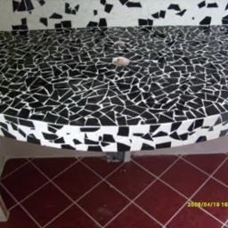 Czarno-biała mozaika z nieregularnych płytek na blacie umywalki w łazience, z widoczną podłogą w brązową kratkę i fragmentem ściany z mozaiką w tle.