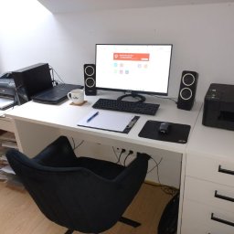 UBEZPIECZENIA GRZEGORZ CHWIAŁKOWSKI - Domowe biuro pod skosem: białe biurko z komputerem, drukarkami, głośnikami i czarnym fotelem obrotowym.
