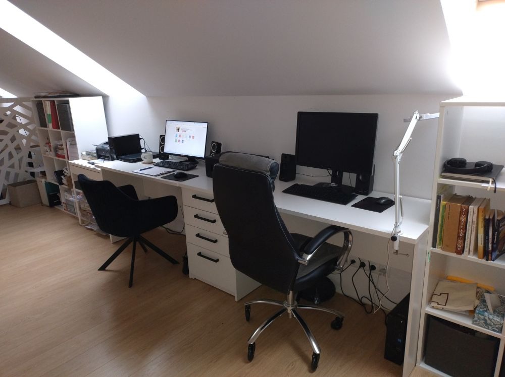 Dwa białe biurka w jasnym pokoju na poddaszu, każde z komputerem, krzesłem biurowym i lampką, półki z książkami w tle.