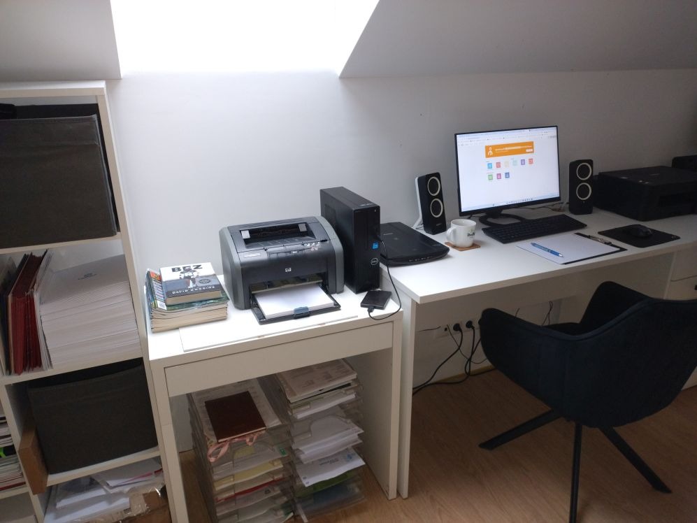 Domowe biuro pod skosem z białym biurkiem, komputerem, drukarką HP, czarnym krzesłem obrotowym i półkami z dokumentami.