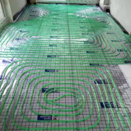 Zielone rury ogrzewania podłogowego rozłożone na izolacji z nadrukiem siatki, widok z góry na dużą powierzchnię, okno w tle.