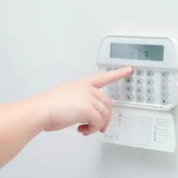 Dłoń z pierścionkiem wciska przycisk '2' na białym panelu sterowania alarmem domowym z wyświetlaczem LCD pokazującym '853'.