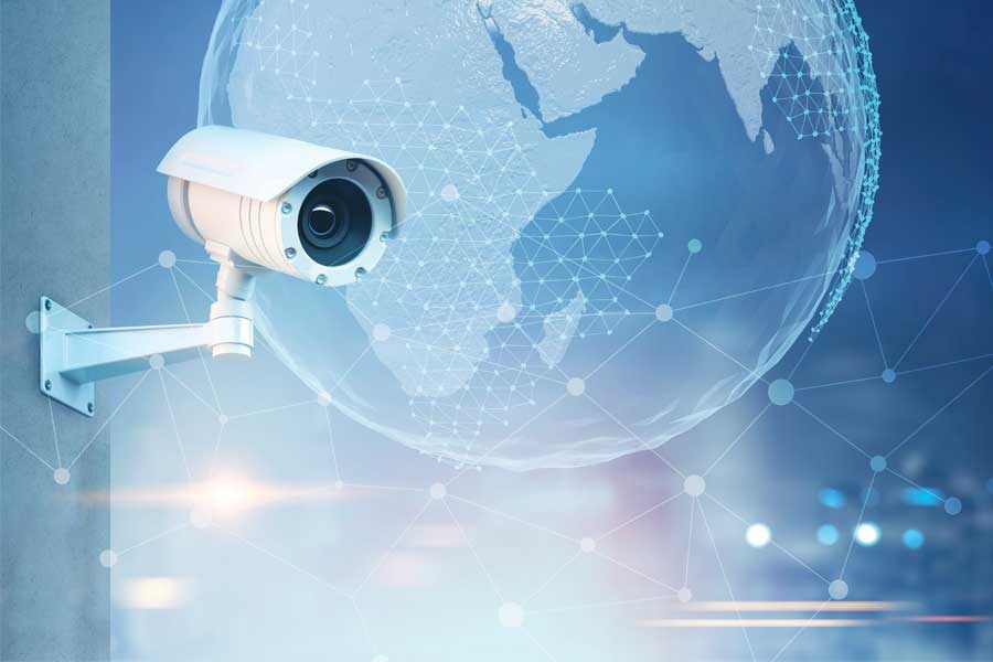 Kamera monitoringu na ścianie z cyfrową nakładką kuli ziemskiej i siatką połączeń, symbolizująca globalny zasięg monitoringu.
