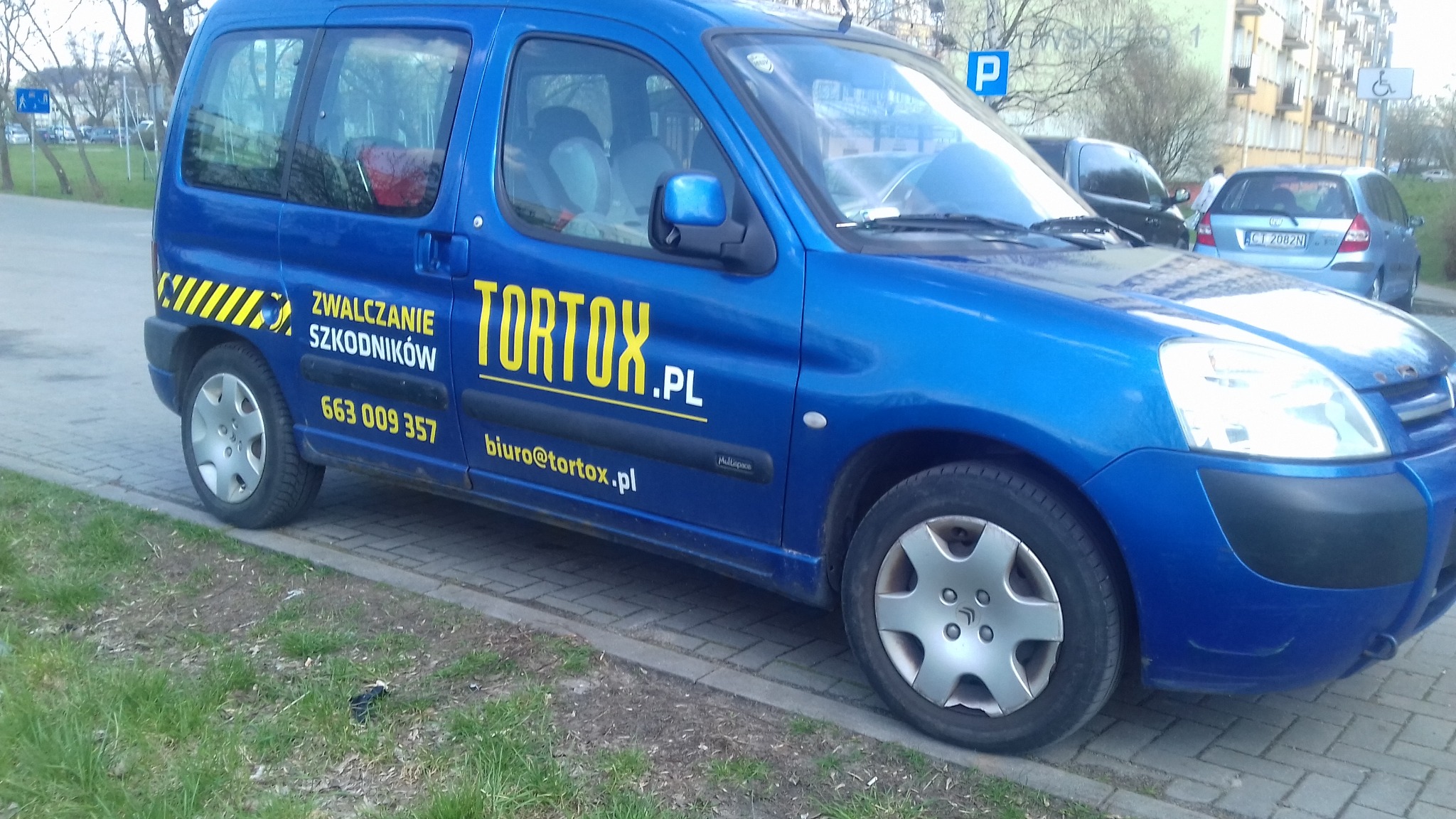 Niebieski samochód firmy Tortox.pl z żółtym napisem 'Zwalczanie szkodników' i numerem telefonu, zaparkowany na chodniku obok trawnika.