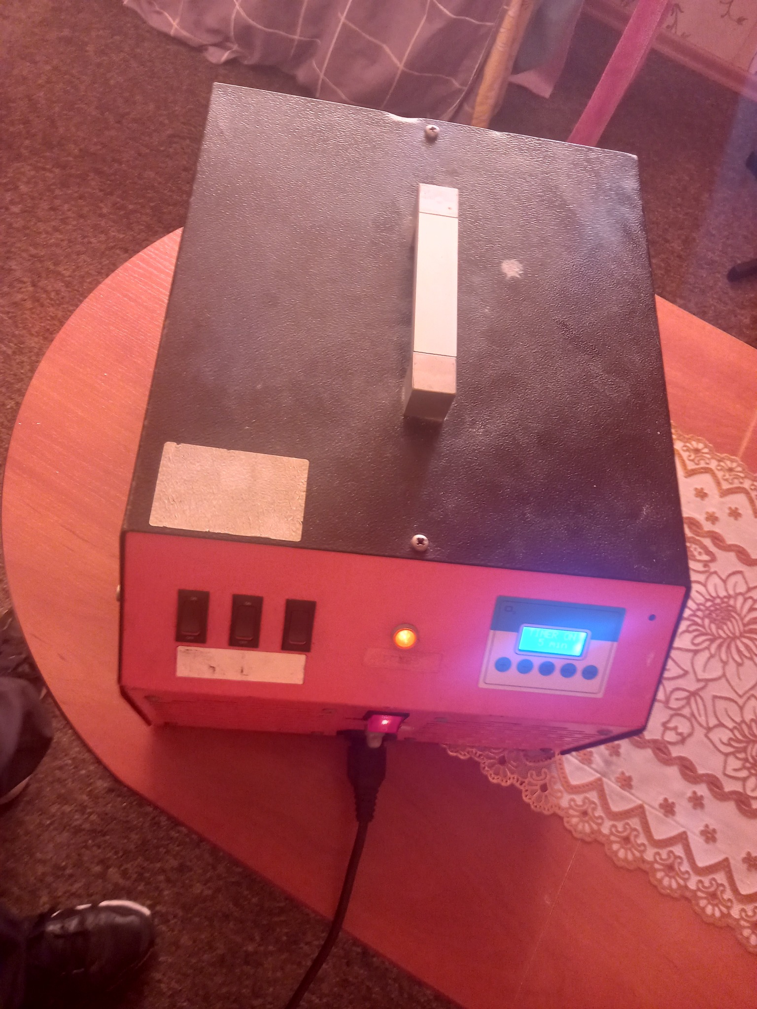 Sprzęt z włączonym wyświetlaczem i trzema przełącznikami, stojący na okrągłym stoliku z koronkową serwetą, podłączony kablem do zasilania.