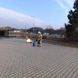 Strażak w kombinezonie ćwiczy gaszenie płomieni na gazie propan-butan podczas szkolenia BHP na zewnątrz. Widoczne butle, gaśnice i kostka brukowa.