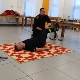 Instruktor w czarnym mundurze demonstruje technikę pierwszej pomocy na manekinie ułożonym na kocu w kratę, w sali z drewnianymi stołami. Kurs pierwszej pomocy.