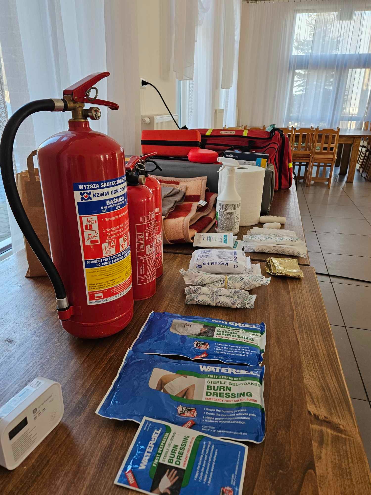 Zestaw BHP na drewnianym stole: gaśnice, apteczka, opatrunki WaterJel na oparzenia, bandaże, koc termiczny, środek dezynfekujący, czujnik czadu. Sprzęt gotowy do użycia.