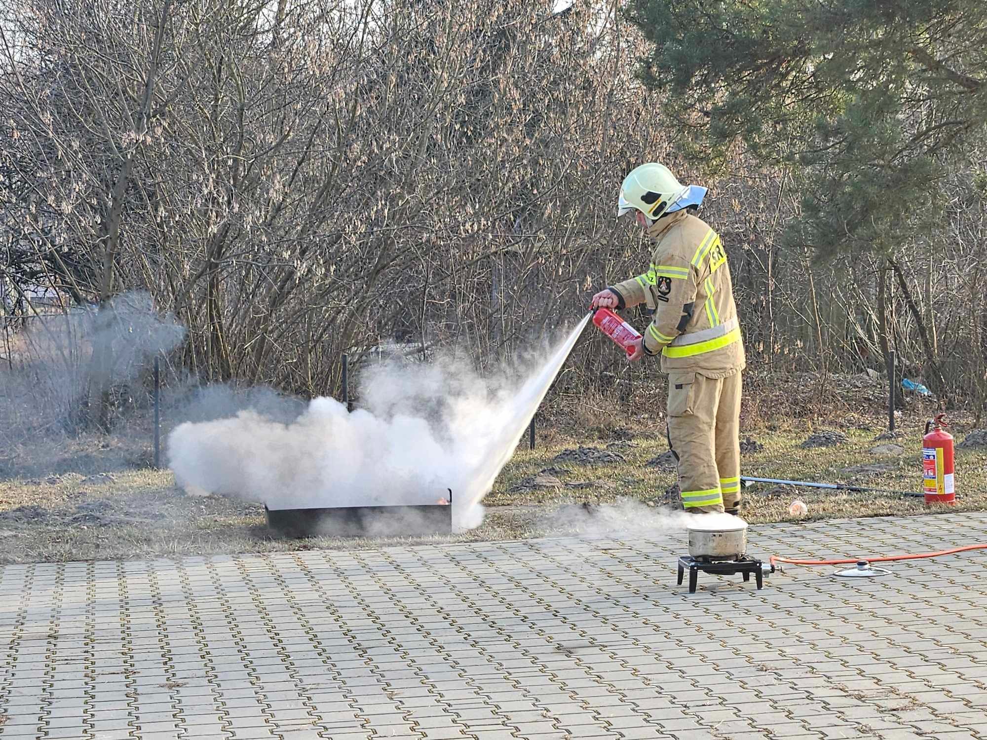 Strażak w hełmie gasi płomień w metalowej skrzyni za pomocą gaśnicy podczas pokazu gaszenia ognia na brukowanym placu, w tle drzewa bez liści i druga gaśnica.
