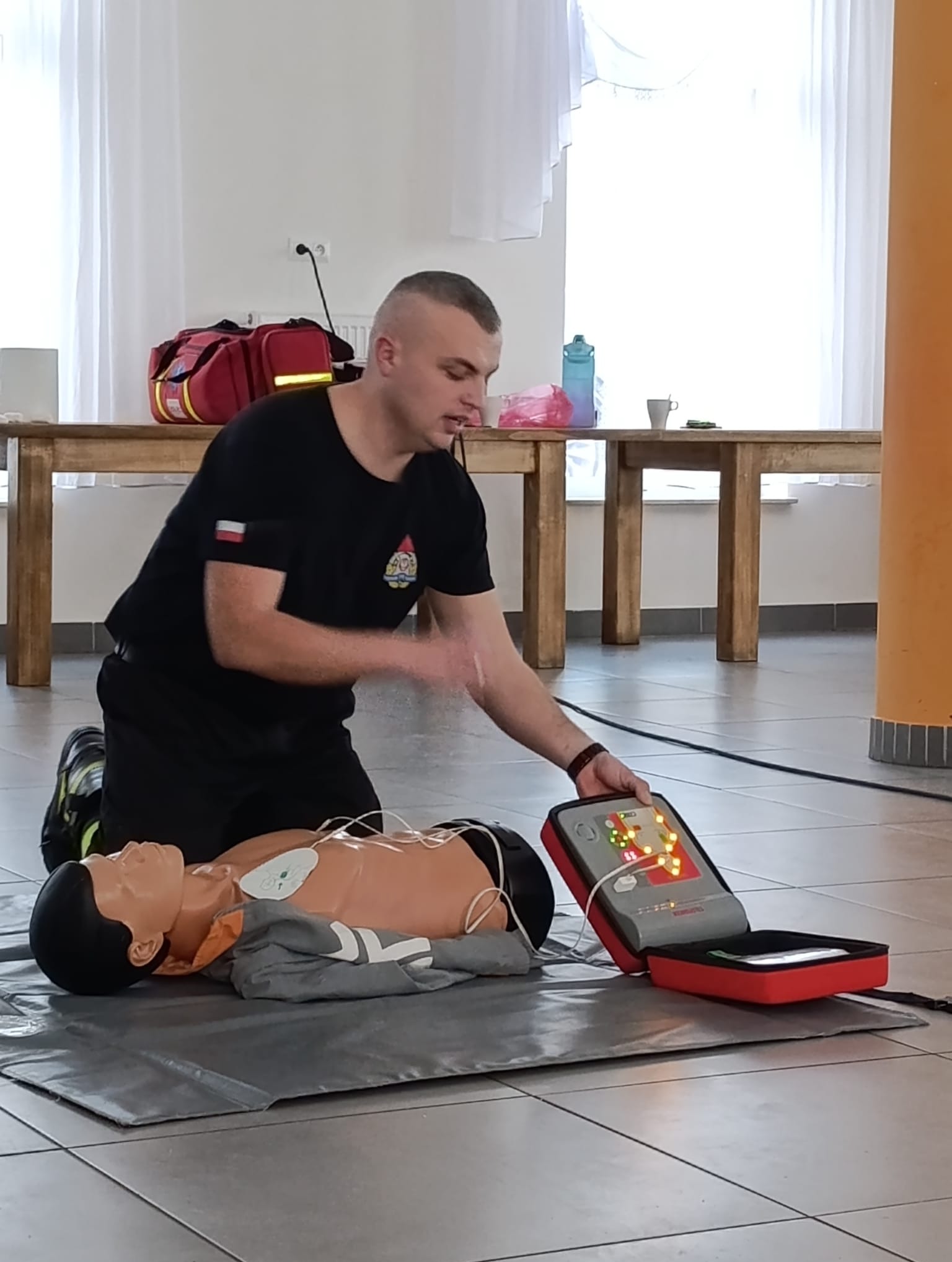 Instruktor demonstruje użycie defibrylatora na fantomie podczas kursu pierwszej pomocy. Widoczny manekin, defibrylator i instruktor w czarnym uniformie.
