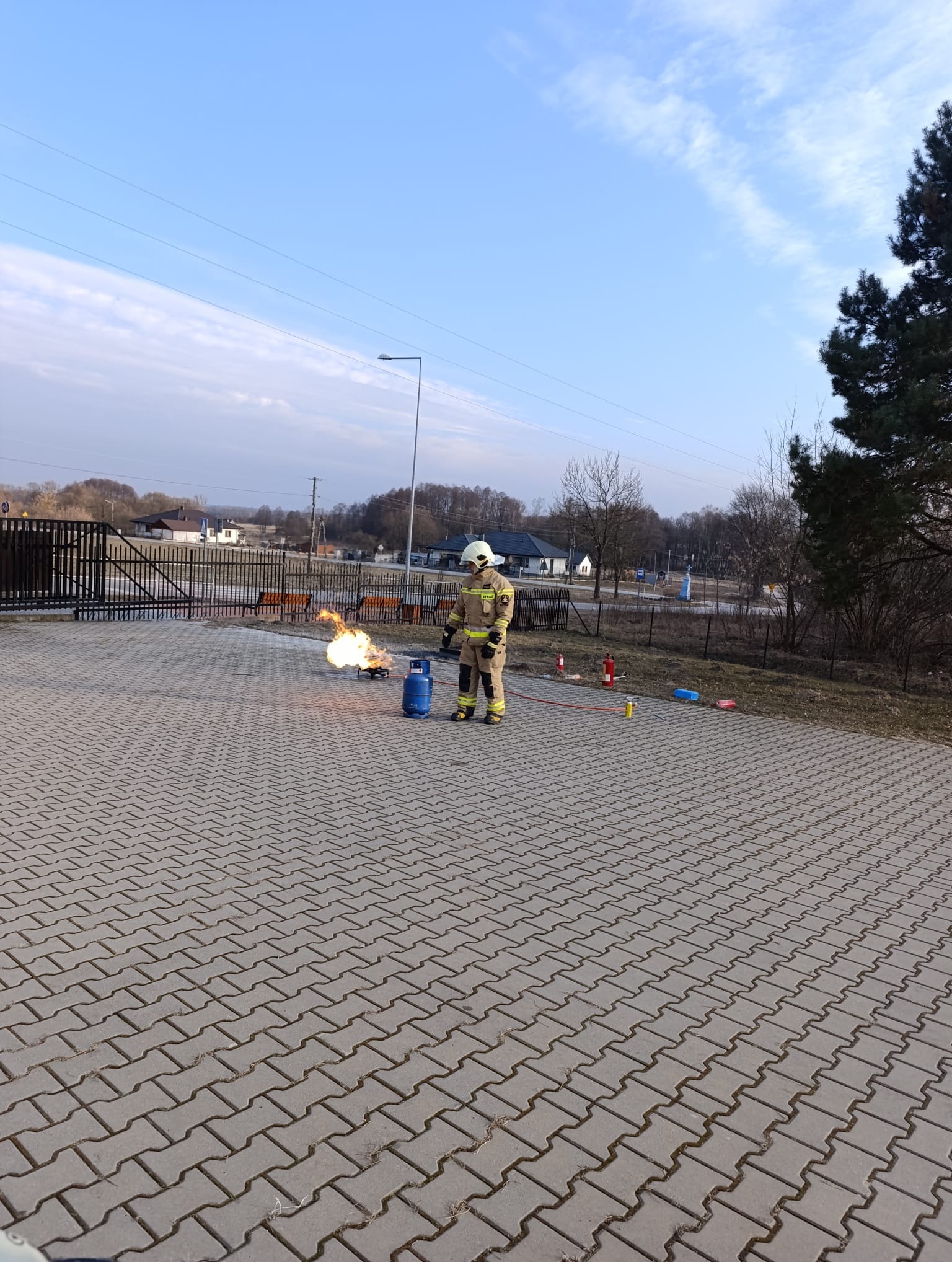 Strażak w kombinezonie ćwiczy gaszenie płomieni na gazie propan-butan podczas szkolenia BHP na zewnątrz. Widoczne butle, gaśnice i kostka brukowa.