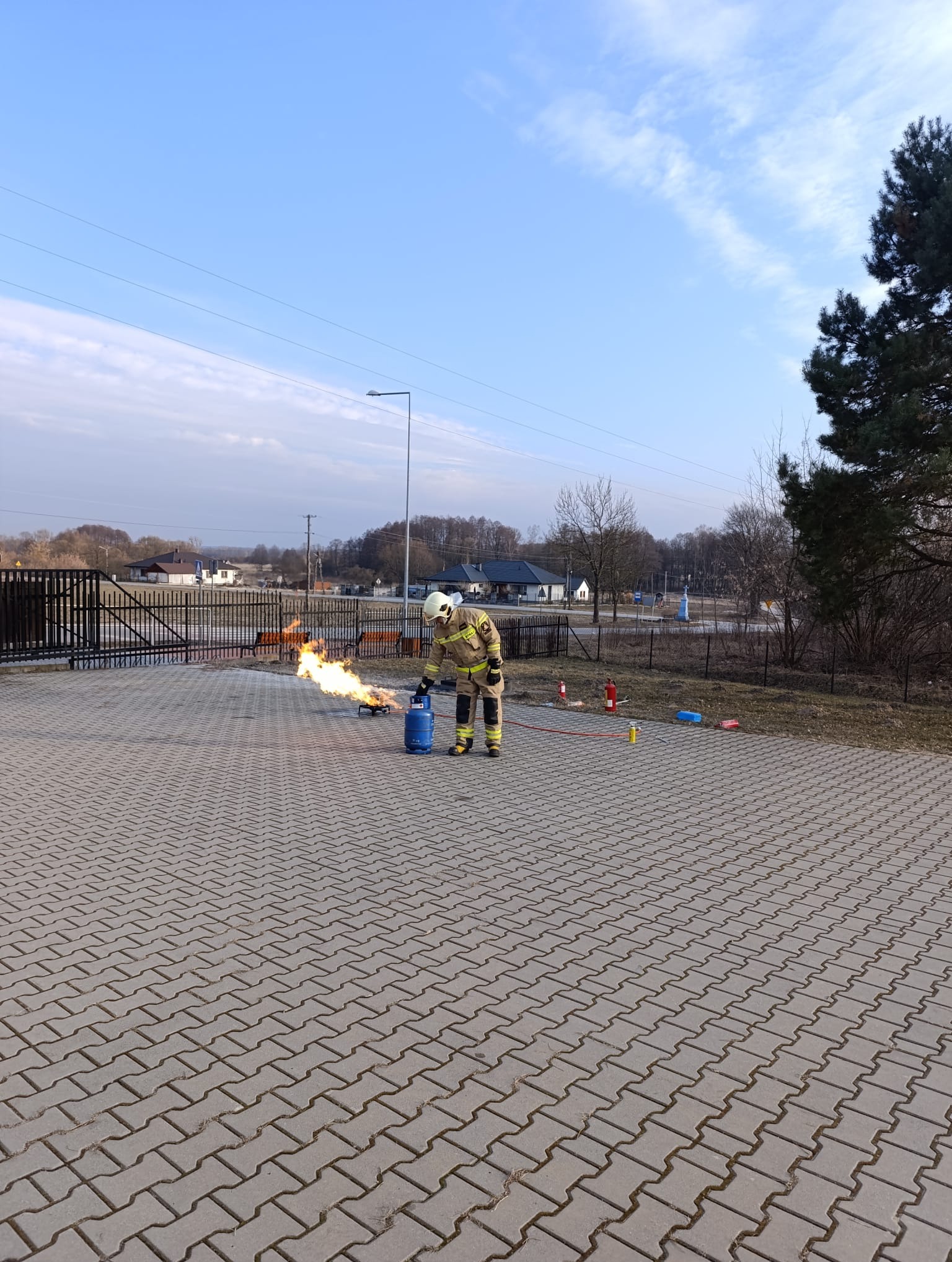 Strażak w kombinezonie demonstruje gaszenie pożaru z butli gazowej podczas pokazu, na tle brukowanej powierzchni i zabudowań.