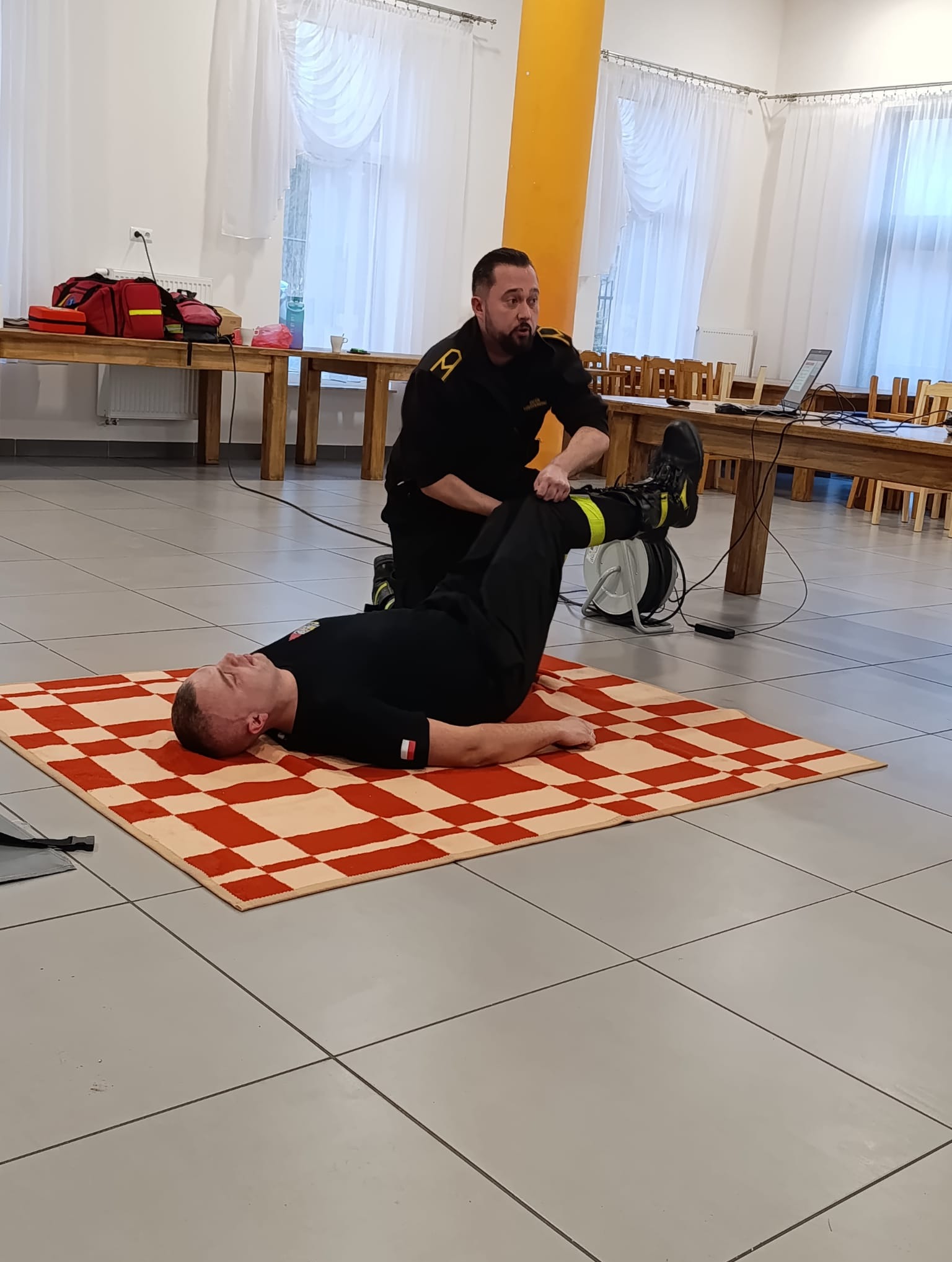 Instruktor w czarnym mundurze demonstruje technikę pierwszej pomocy na manekinie ułożonym na kocu w kratę, w sali z drewnianymi stołami. Kurs pierwszej pomocy.