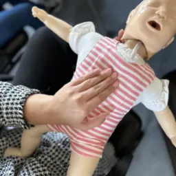 Instruktor z zadbanymi paznokciami demonstruje uciskanie klatki piersiowej na manekinie niemowlęcia ubranego w biało-różowe paski.