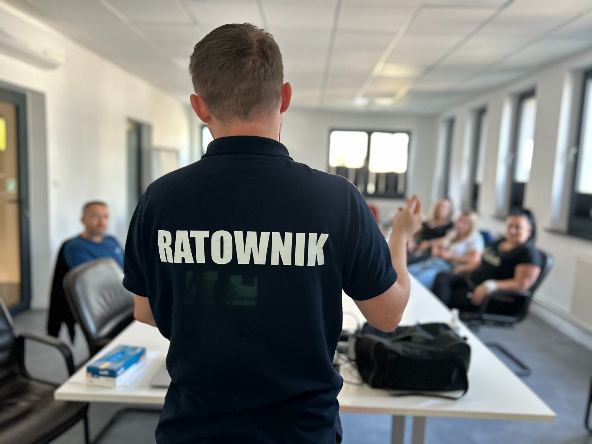 Prelegent z napisem 'RATOWNIK' na koszulce prowadzi zajęcia dla grupy osób siedzących przy stole w jasnej sali konferencyjnej.