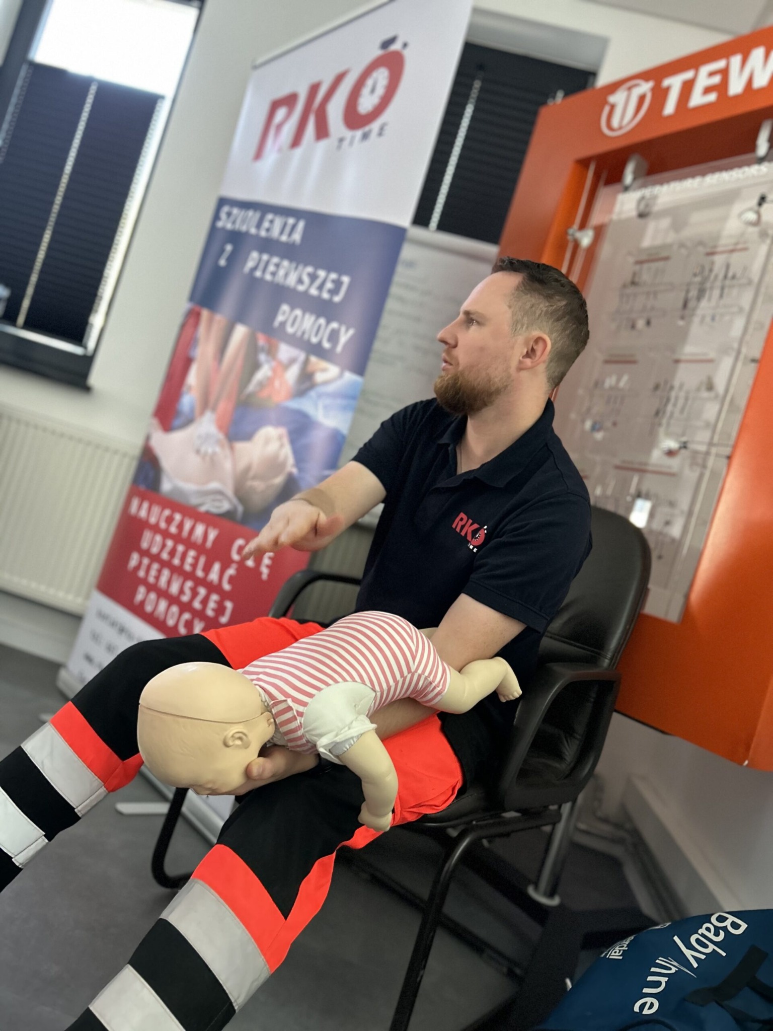 Instruktor w stroju ratownika medycznego demonstruje resuscytację krążeniowo-oddechową na manekinie niemowlęcia podczas szkolenia z pierwszej pomocy, w tle baner informacyjny i sprzęt dydaktyczny.