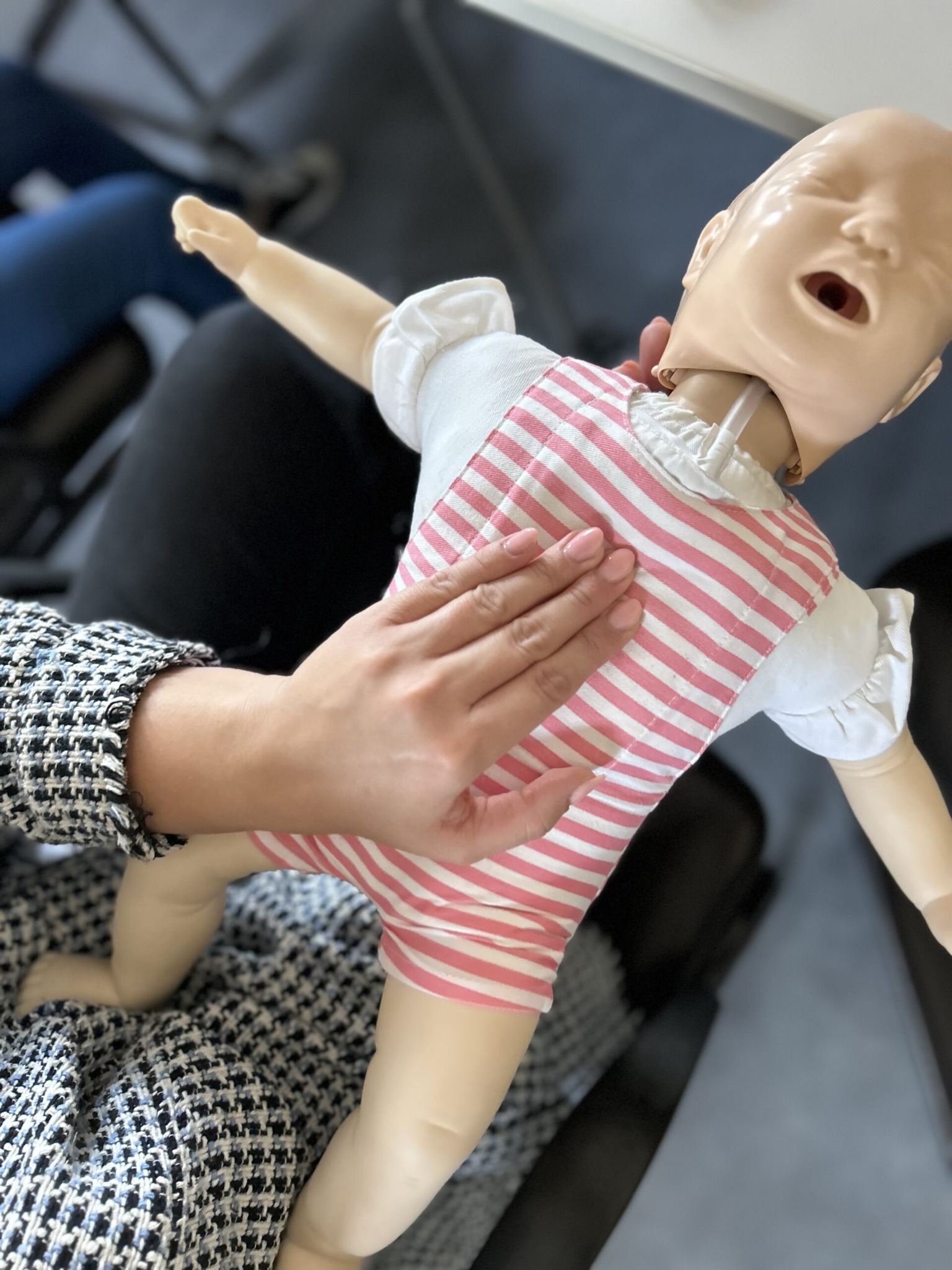 Instruktor z zadbanymi paznokciami demonstruje uciskanie klatki piersiowej na manekinie niemowlęcia ubranego w biało-różowe paski.