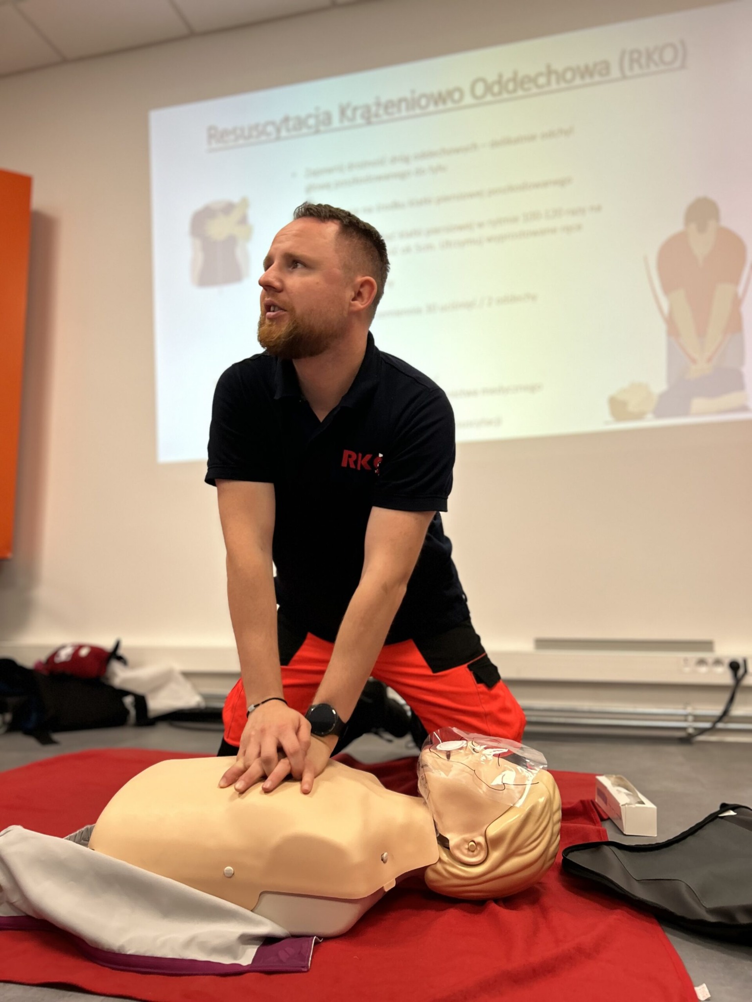 Instruktor w czarnej koszulce z czerwonym logo RKS demonstruje resuscytację krążeniowo-oddechową na manekinie podczas szkolenia, w tle widoczny rzutnik z prezentacją RKO.