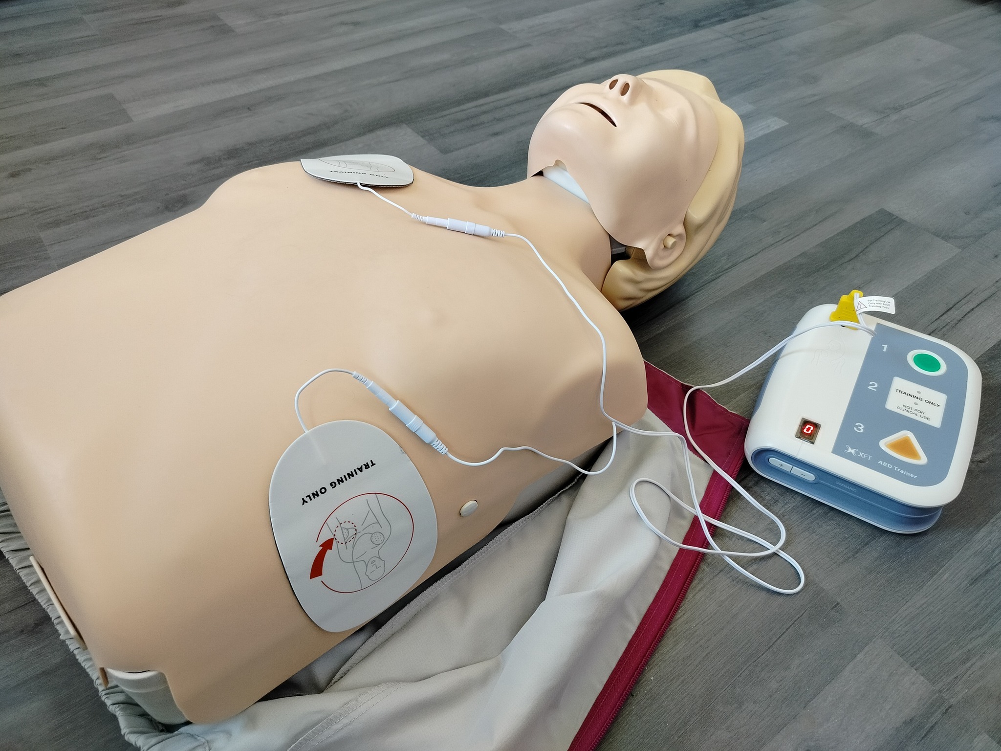 Manekin do ćwiczeń resuscytacji krążeniowo-oddechowej z podłączonym defibrylatorem treningowym w Lublinie, leżący na szarym panelu podłogowym. Na manekinie widoczne elektrody treningowe.