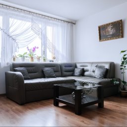 DomVisio - Szary narożnik z poduszkami, czarny stolik kawowy i rośliny doniczkowe w jasnym salonie z parkietem, firankami i obrazem w złotej ramie.