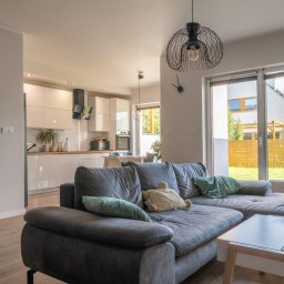 DomVisio - Jasne wnętrze domu z szarym narożnikiem, widok na ogród przez duże okno, otwarta kuchnia w tle, lampa druciana nad narożnikiem.