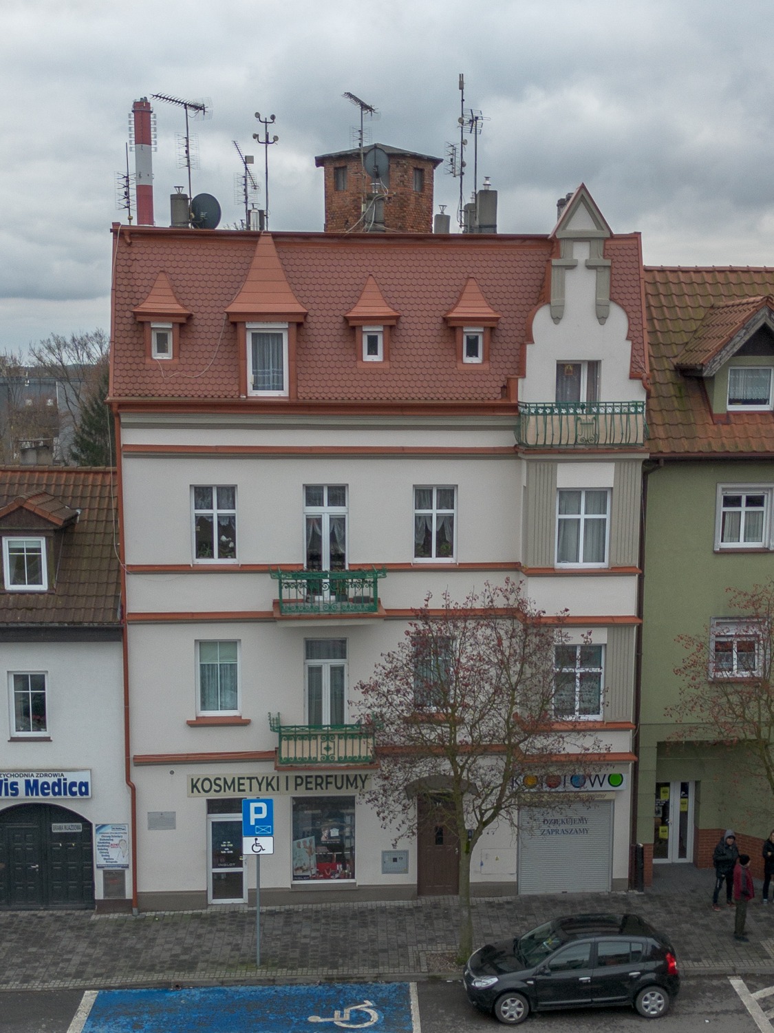 Trzypiętrowy budynek z balkonami i sklepem 'Kosmetyki i Perfumy' na parterze, widok z góry na parking dla niepełnosprawnych i zaparkowany czarny samochód.
