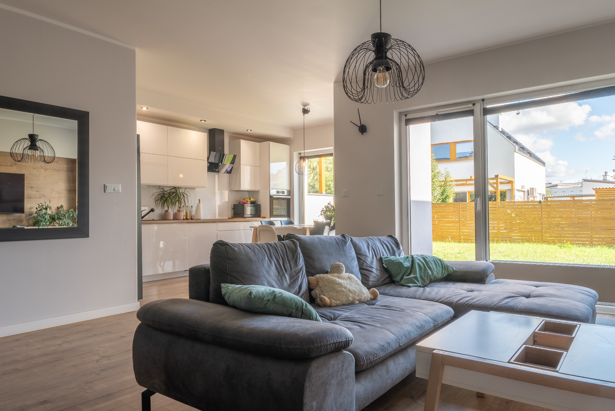 Jasne wnętrze domu z szarym narożnikiem, widok na ogród przez duże okno, otwarta kuchnia w tle, lampa druciana nad narożnikiem.