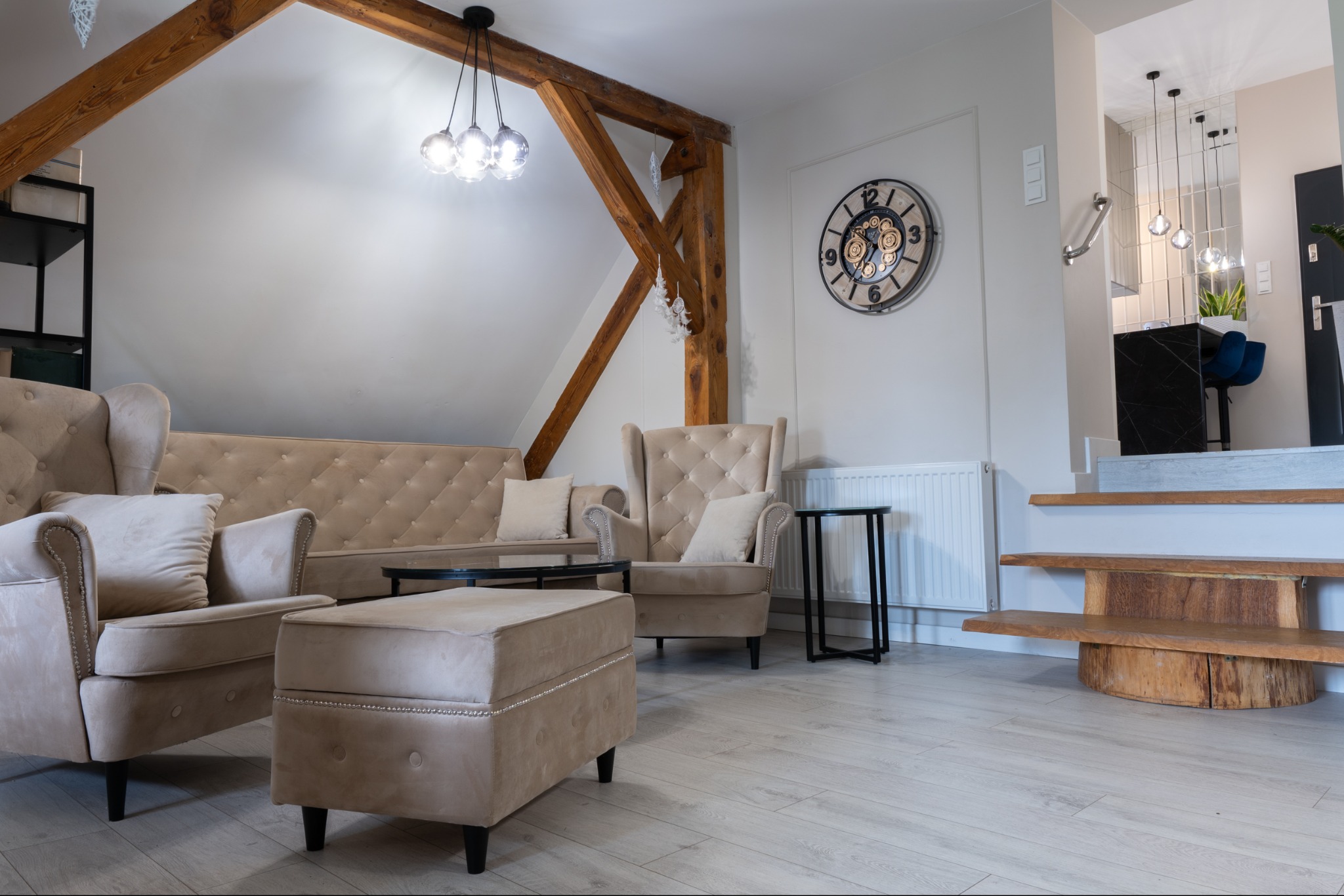 Jasne wnętrze salonu z beżowymi meblami wypoczynkowymi, drewnianą belką stropową i dekoracyjnym zegarem na ścianie, schody z drewnianymi stopniami prowadzące do kuchni.