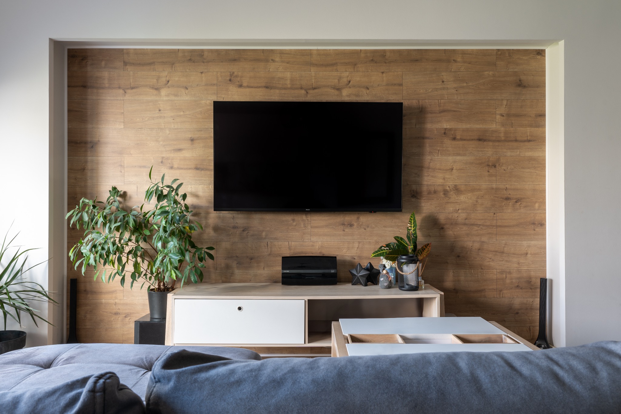 Telewizor zawieszony na drewnianej ścianie za jasnym, nowoczesnym stolikiem RTV z roślinami doniczkowymi i dekoracjami, widok zza szarej kanapy.