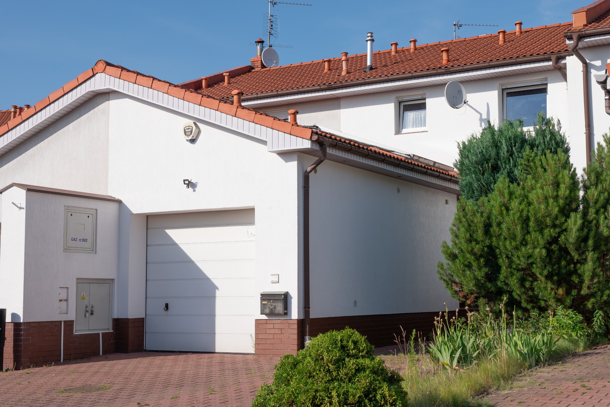 Biały dom szeregowy z garażem, pomarańczową dachówką i brązowymi rynnami, widoczny z brukowanego podjazdu.