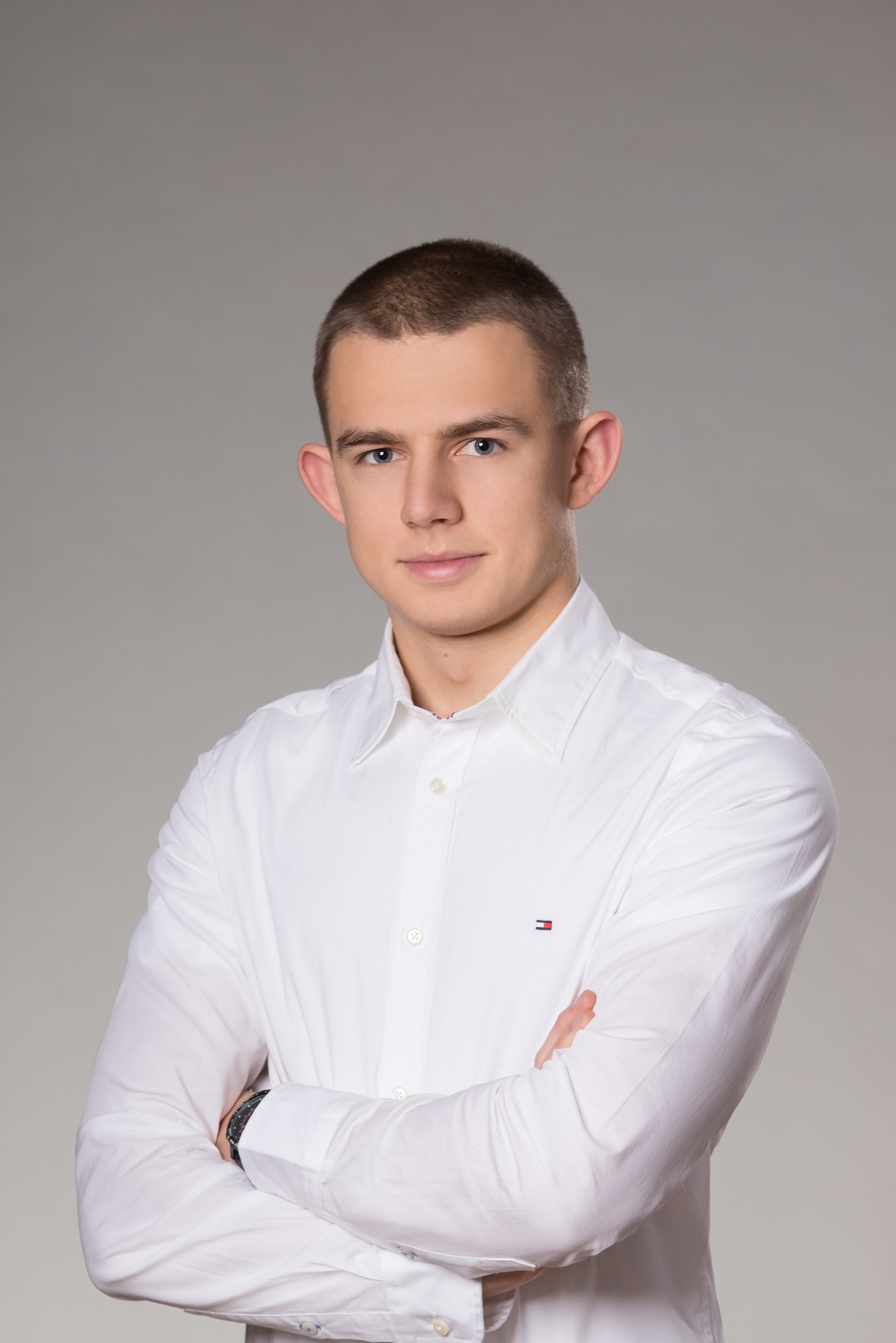 Portret młodego mężczyzny w białej koszuli z logo Tommy Hilfiger, skrzyżowane ręce, zegarek na lewym nadgarstku, na jednolitym szarym tle.