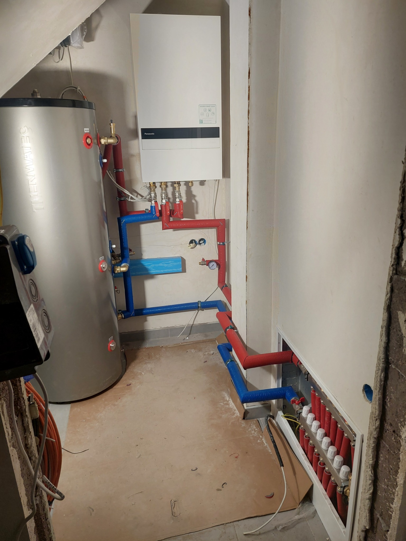 Bojler, piec gazowy i rozdzielacz ogrzewania podłogowego z rurami w kolorze czerwonym i niebieskim, zamontowane w pomieszczeniu w trakcie remontu.