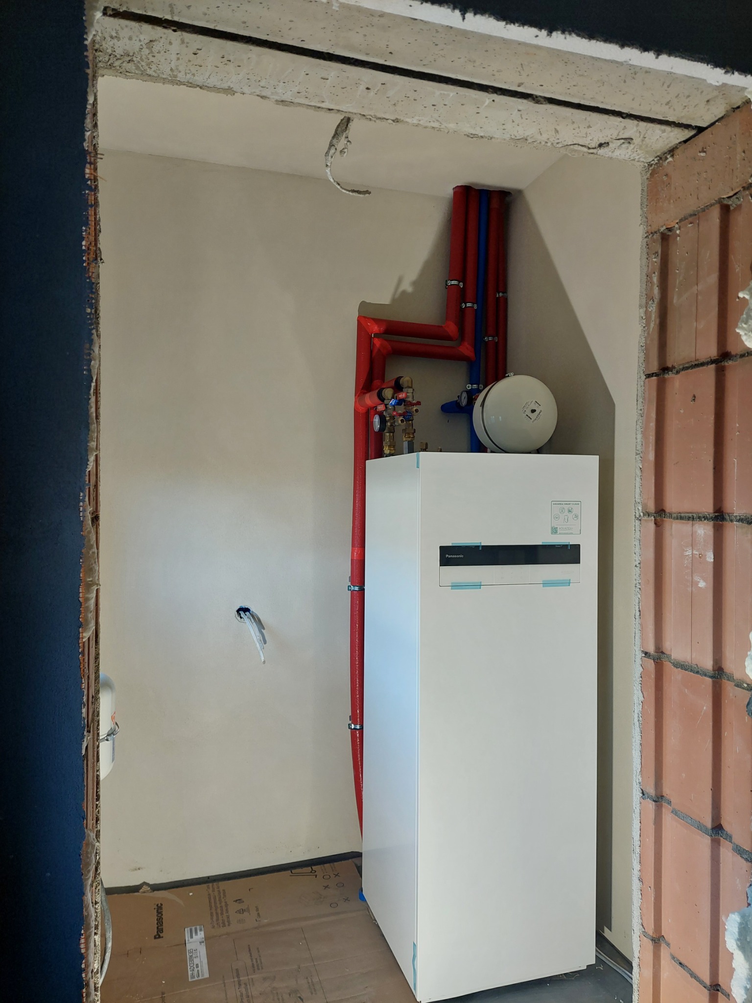 Nowy biały piec gazowy Panasonic z czerwonymi i niebieskimi rurami podłączony w surowym pomieszczeniu z ceglaną ścianą i odsłoniętym betonem.
