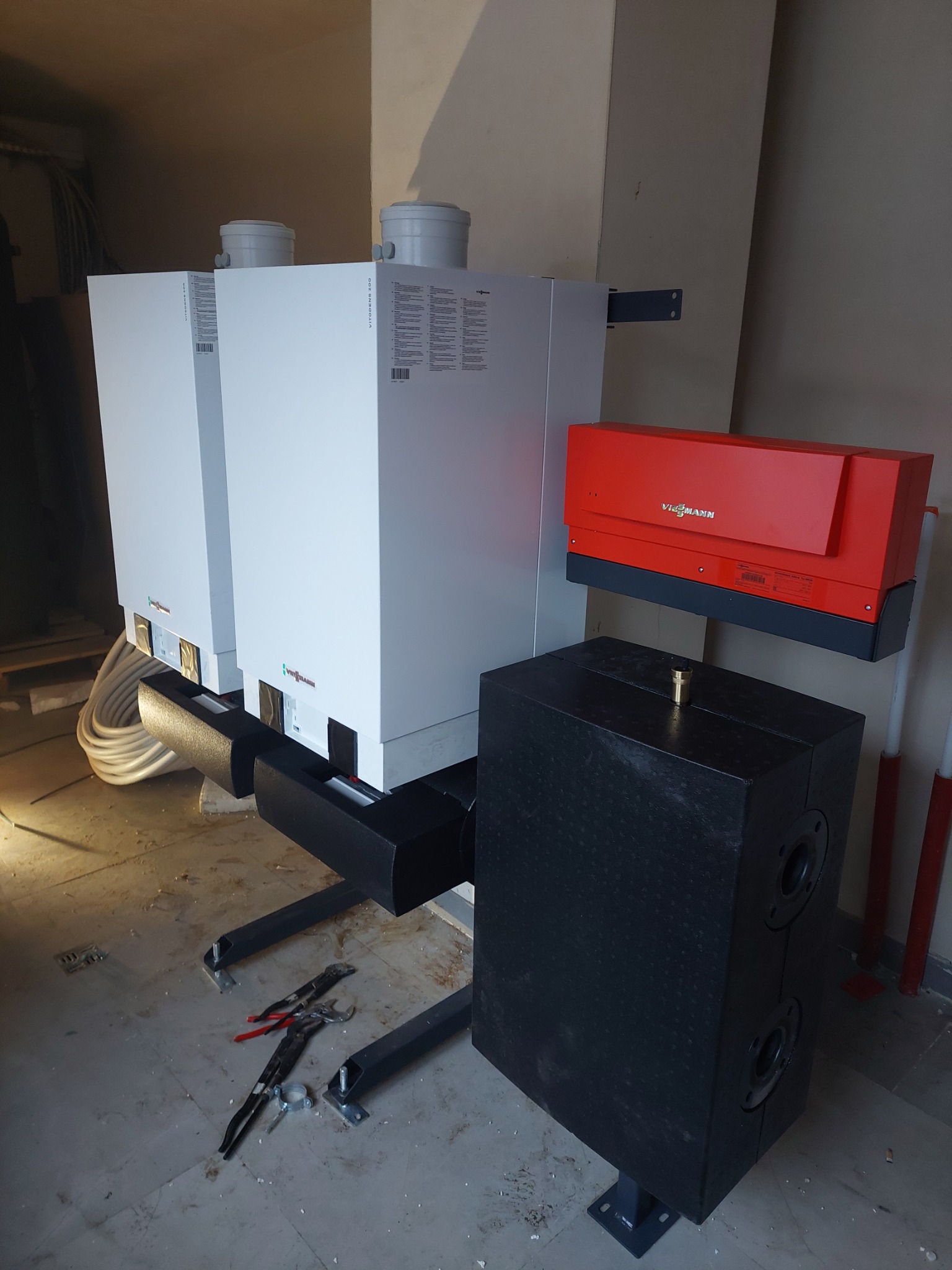 Dwa białe piece gazowe Viessmann Vitodens 200 z czerwonym regulatorem, zamontowane na czarnej konstrukcji stalowej, obok czarny zasobnik, na podłodze leżą narzędzia hydrauliczne.