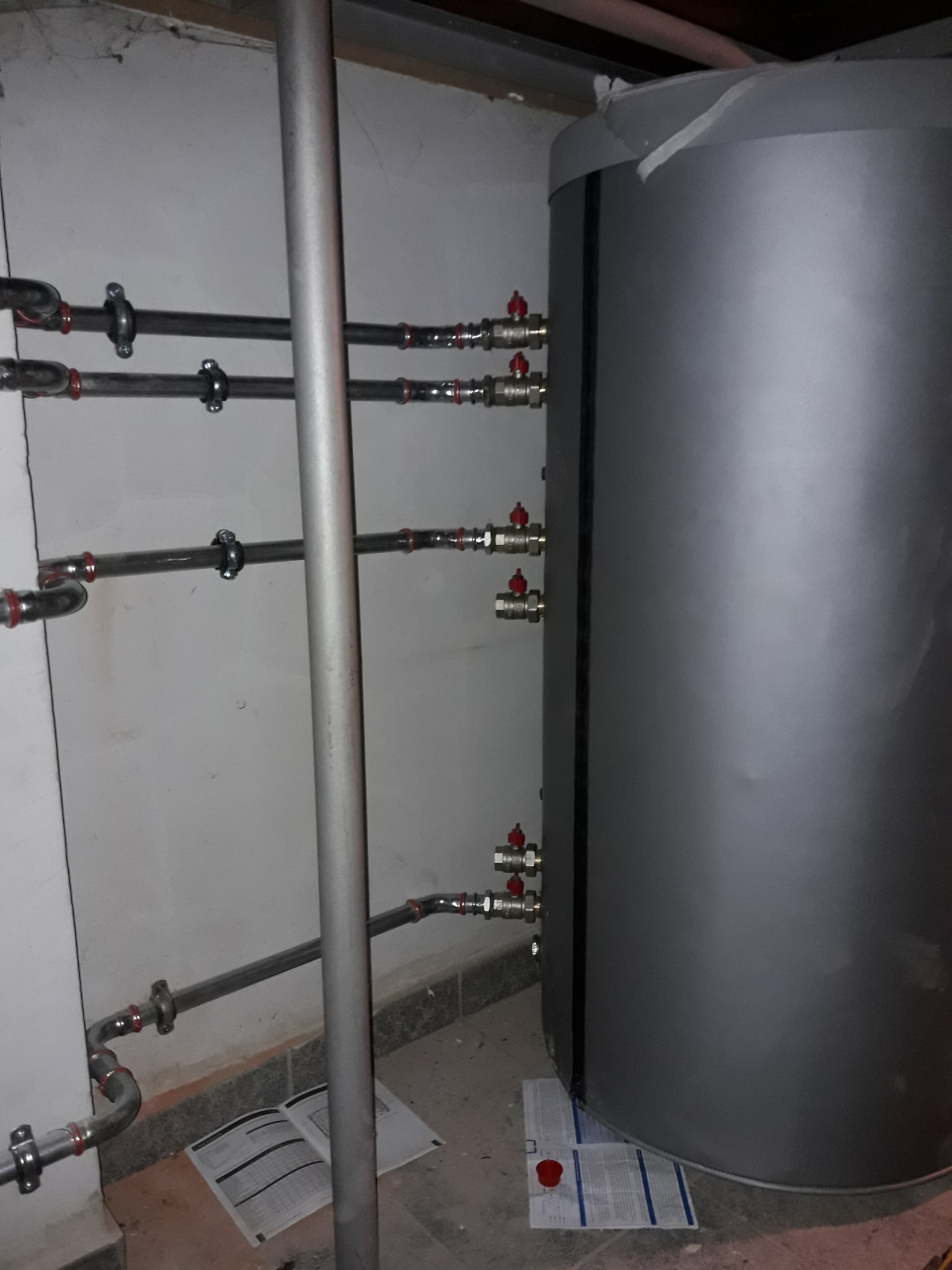 Szary bojler z podłączonymi rurami i zaworami z czerwonymi pokrętłami, widoczne fragmenty instalacji hydraulicznej oraz instrukcja obsługi na podłodze