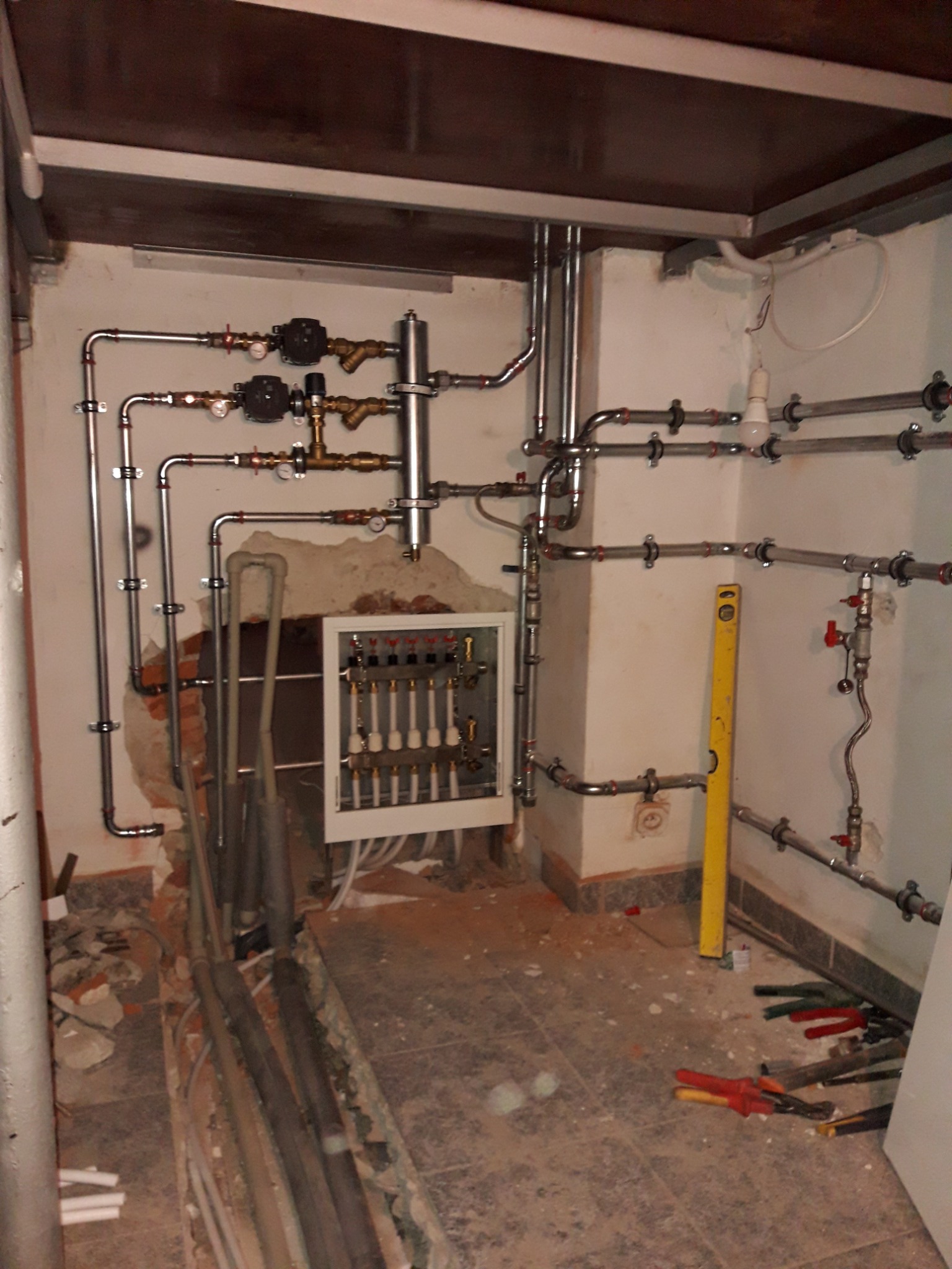 Rozbudowana instalacja hydrauliczna i grzewcza w piwnicy, widoczne rury miedziane, zawory, rozdzielacz w szafce, poziomica, narzędzia hydrauliczne na podłodze w trakcie prac wykończeniowych.