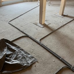 ELEKTRODA Marcin Kluska - Wnętrze w trakcie remontu z widocznymi przewodami elektrycznymi ułożonymi na podłodze, zabezpieczonymi peszlami i przymocowanymi do podłoża klamrami, przykryte częściowo szarą plandeką, w tle...