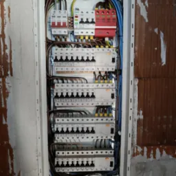 Rozdzielnica elektryczna z zainstalowanymi bezpiecznikami, wyłącznikami różnicowoprądowymi i przewodami, zamontowana w surowej ścianie z cegły.