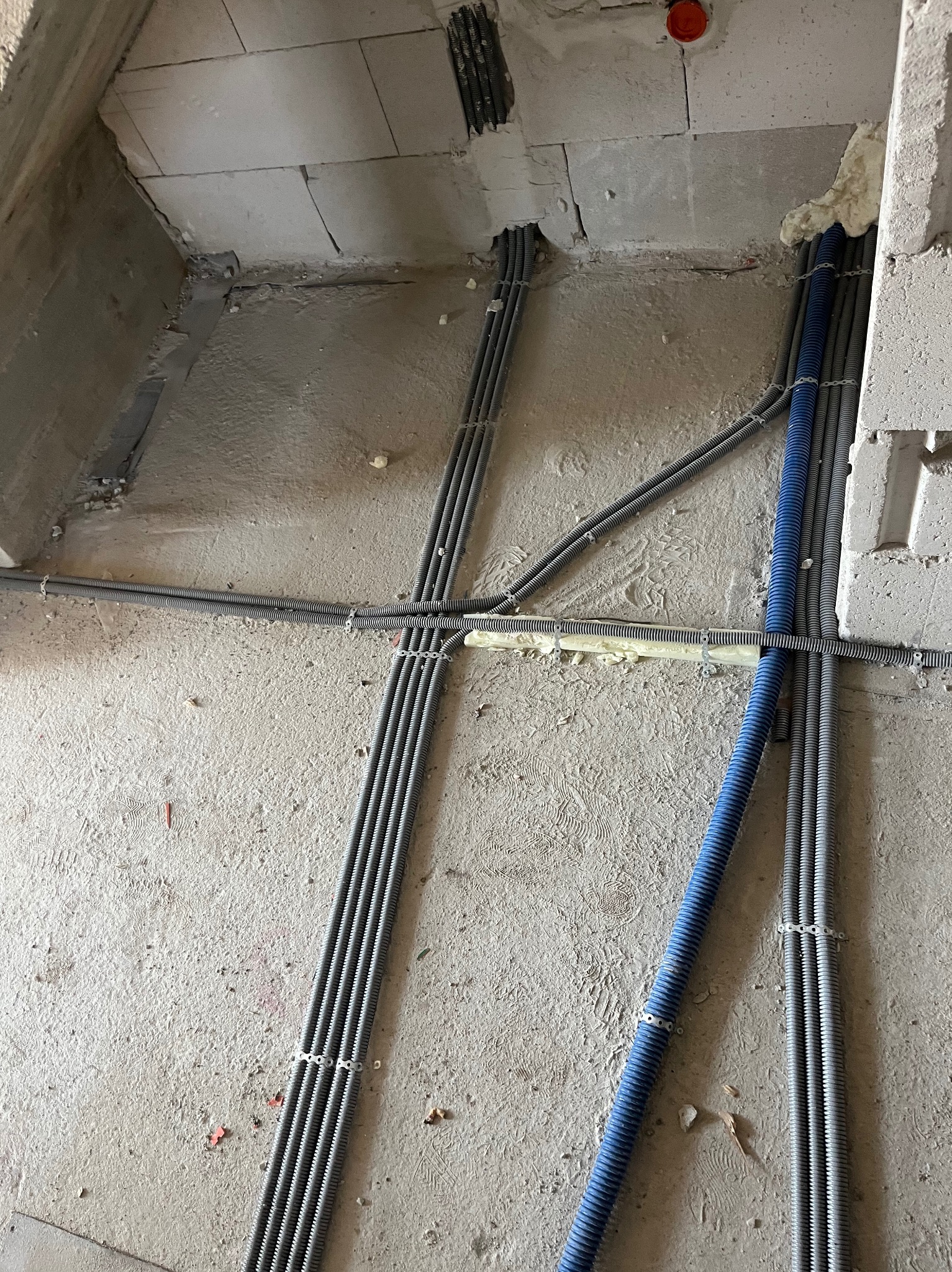 Szare i niebieskie rury karbowane prowadzone po surowej ścianie z betonu komórkowego, zabezpieczone uchwytami i pianką montażową.