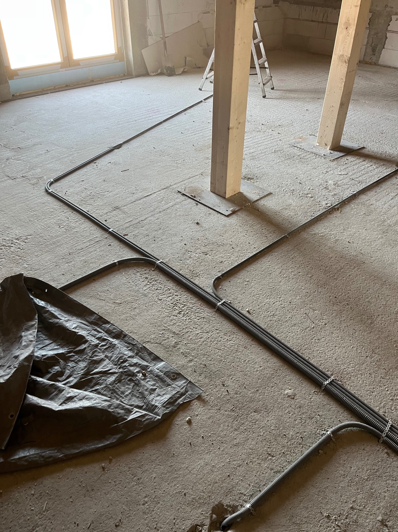 Wnętrze w trakcie remontu z widocznymi przewodami elektrycznymi ułożonymi na podłodze, zabezpieczonymi peszlami i przymocowanymi do podłoża klamrami, przykryte częściowo szarą plandeką, w tle...