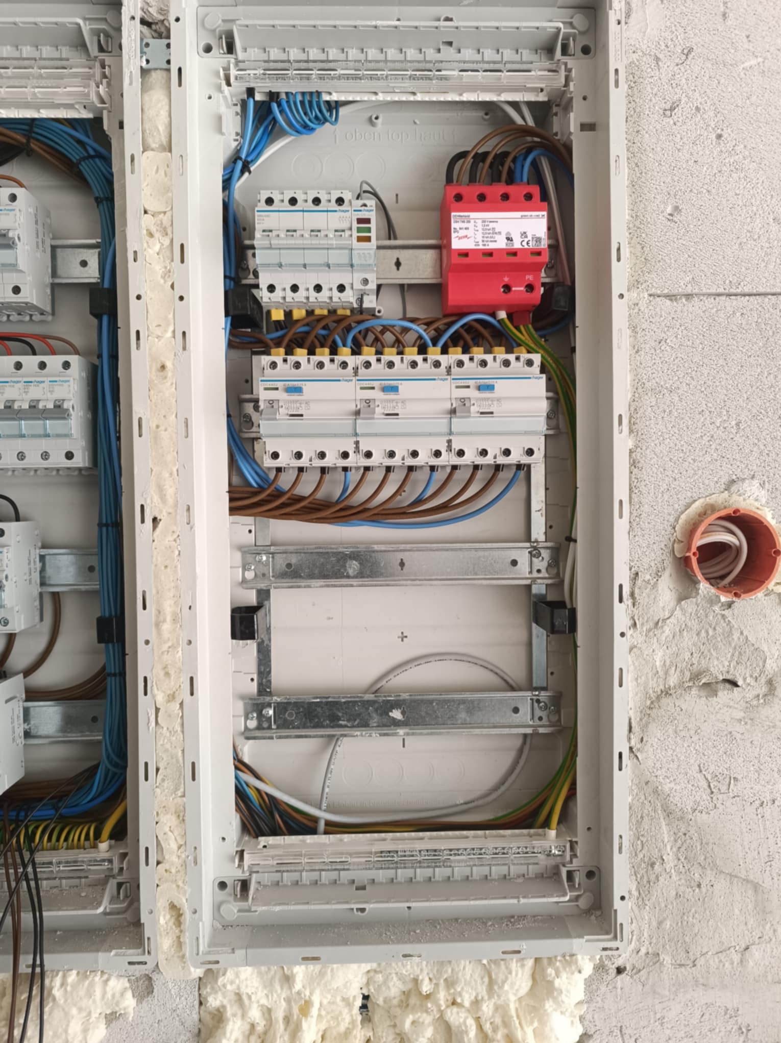 Rozdzielnica elektryczna w trakcie montażu, widoczne zabezpieczenia przepięciowe (czerwony moduł), wyłączniki nadprądowe Hager, okablowanie w kolorach brązowym, niebieskim i zielono-żółtym, ściana...