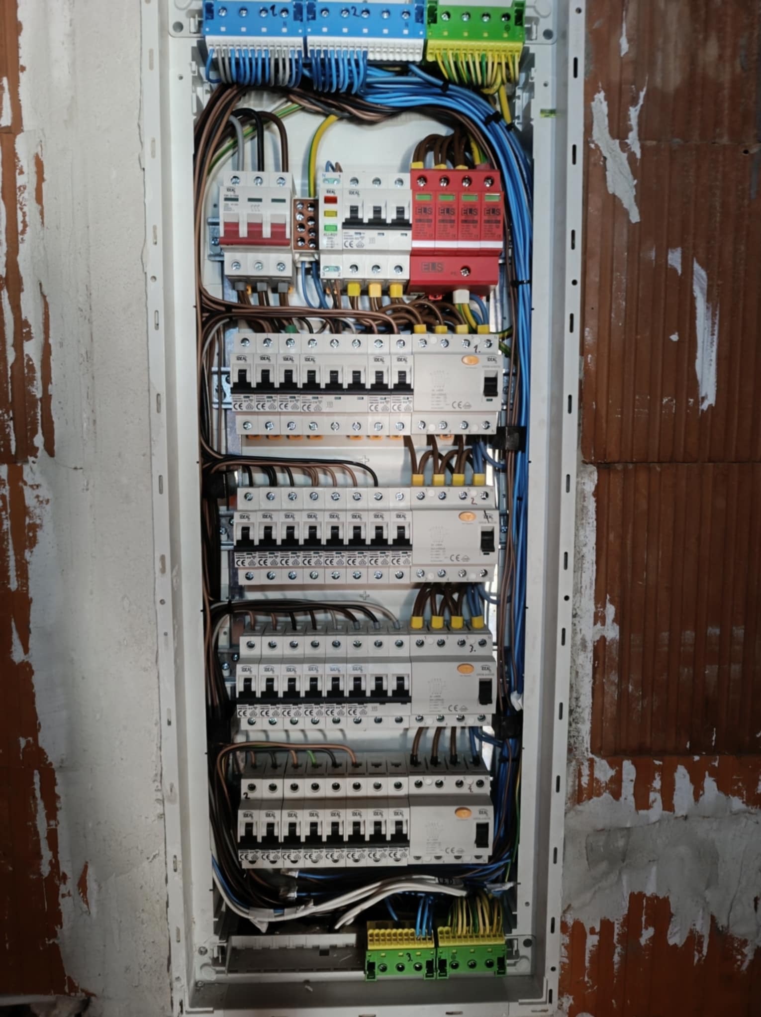 Rozdzielnica elektryczna z zainstalowanymi bezpiecznikami, wyłącznikami różnicowoprądowymi i przewodami, zamontowana w surowej ścianie z cegły.