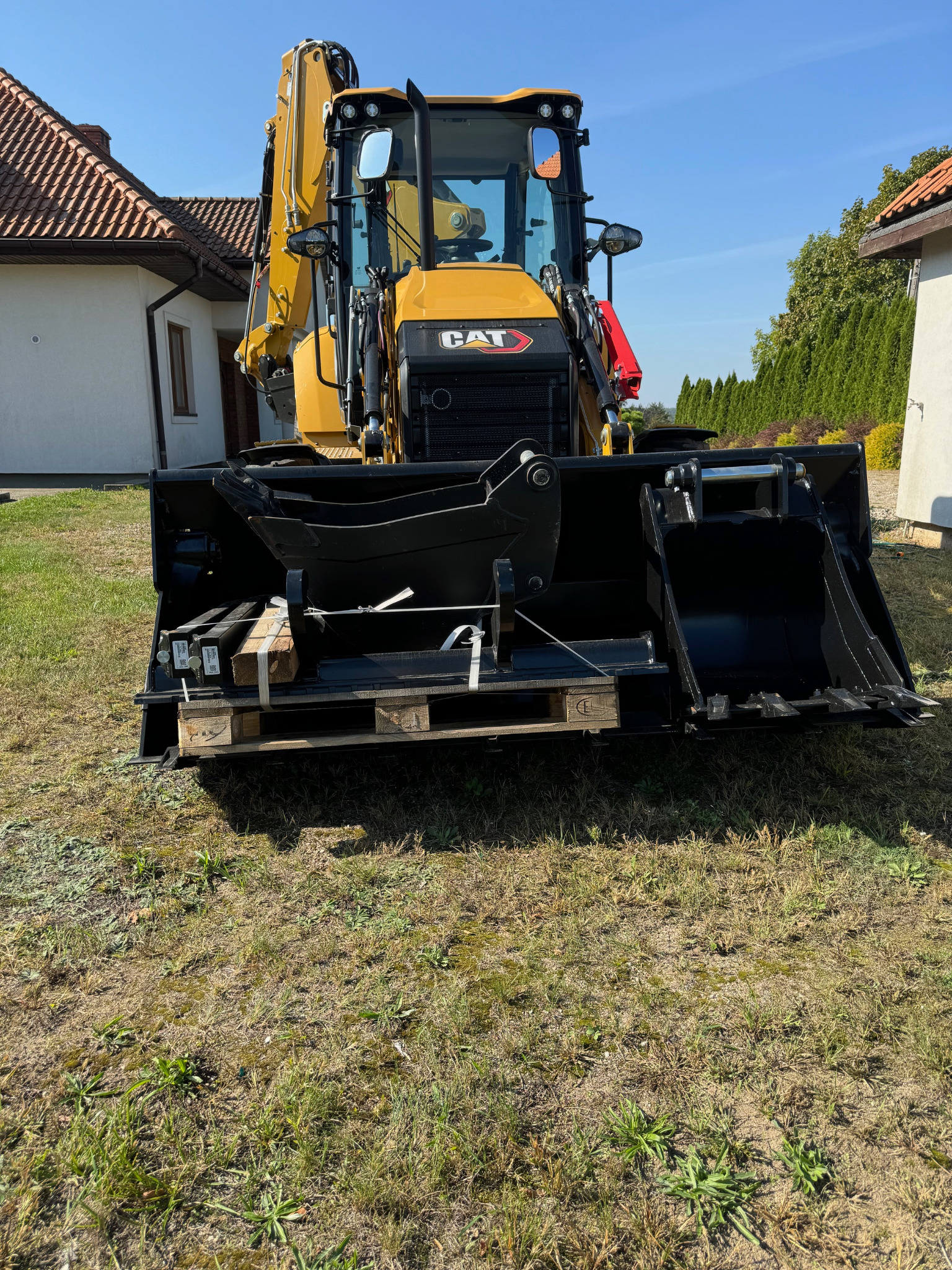 Żółta koparko-ładowarka CAT z czarną łyżką na trawiastym terenie, z paletą i drewnianymi elementami wewnątrz łyżki. W tle dom z czerwoną dachówką i zielone tuje.