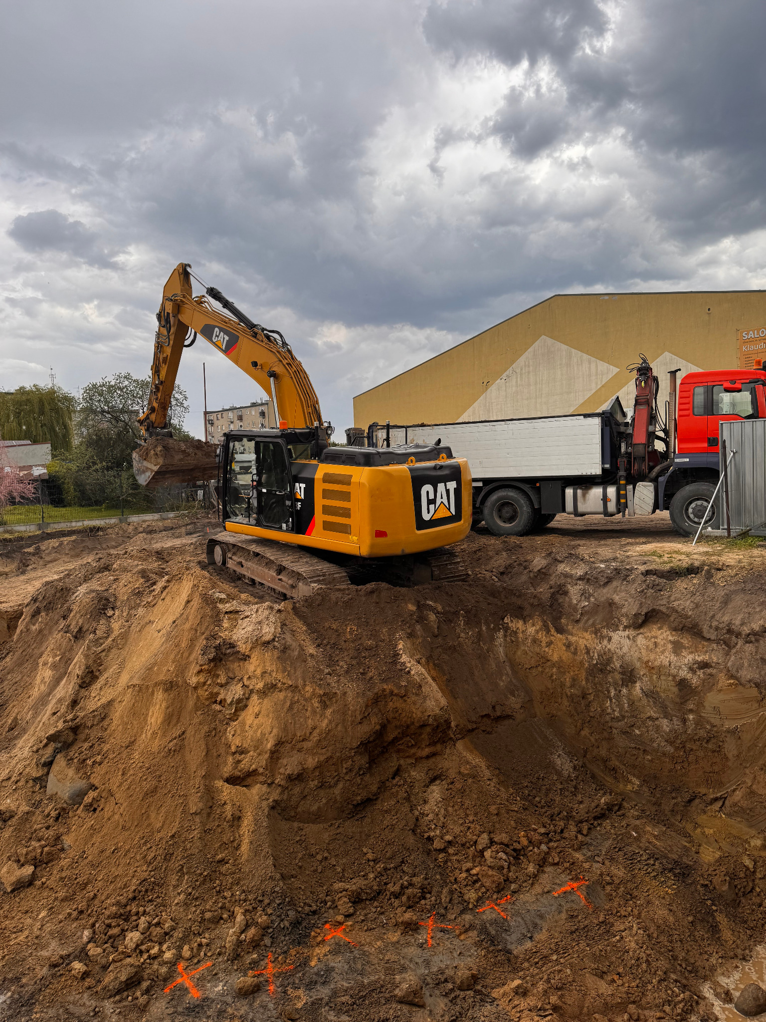 Żółta koparka CAT na placu budowy, wykop pod fundamenty, obok ciężarówka z dźwigiem, pochmurne niebo. Widoczne pomarańczowe oznaczenia w ziemi.
