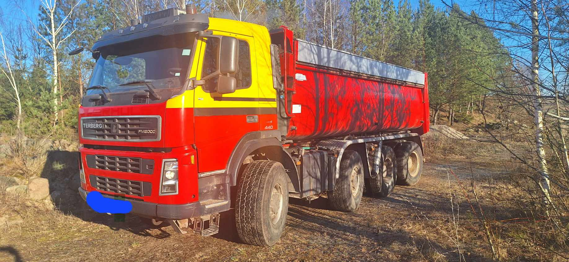 Czerwono-żółta wywrotka Terberg FM2000 na tle drzew, z widoczną stertą ziemi w tle. Pojazd zaparkowany na nierównym terenie.